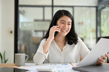 Asian young businesswoman talking on the phone while looking the details on her report.