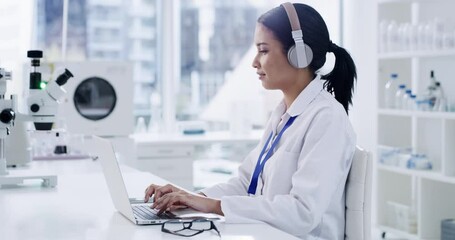 Young female scientist working on laptop with wireless headphones to do research in a modern laboratory. Medical professional browsing the internet. Smart doctor typing on a computer in a bright lab.