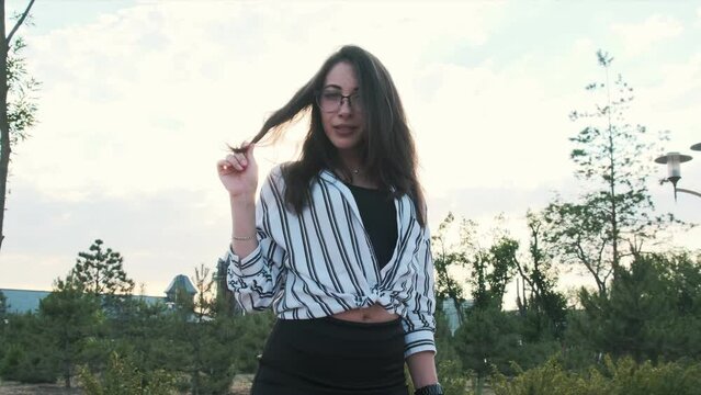 Beautiful girl in black high-heeled shoes, a black skirt and a striped shirt is standing in the park, posing and looking at the camera. Camera zoom, slow motion

