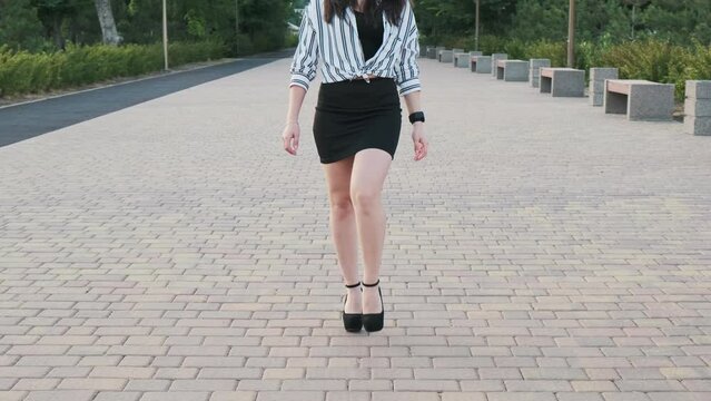 Beautiful girl in black high-heeled shoes, a black skirt and a striped shirt walks in the park. Front view. Slow motion
