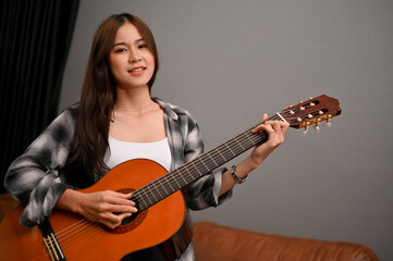 Fototapeta premium Beautiful young Asian female artist playing an acoustic guitar in her home living room.