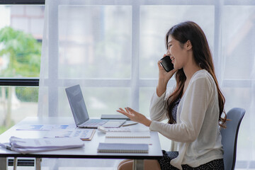 Asian businesswoman using smartphone to do accounting work with laptop and graph of financial reports in office