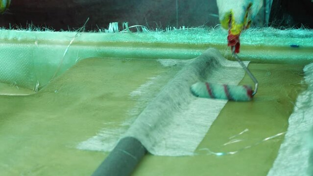 A man's hand with a paint roller applies adhesive to the fiberglass, gluing the hull of a pedal catamaran. Building fiberglass boats in a private shop