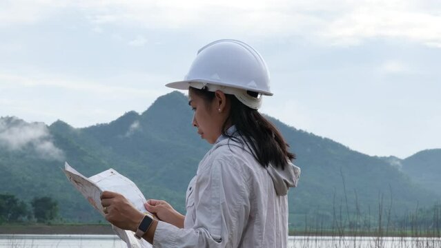 Engineering ecologist woman in a helmet holding a blueprint stands on the bank of a river to develop a hydroelectric dam to generate electricity. Clean energy and Technology concepts.