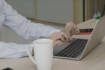 A woman clenched her left hand to exercise her finger from fatigue while typing a laptop on a table with a white coffee mug and a mobile phone next to it. Employees in white shirts sit and print work
