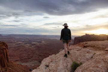 Fototapeta na wymiar Adventurous Woman Hiking at a Desert Canyon with Red Rock Mountains. Cloudy Sunset Sky. Canyonlands National Park. Utah, United States. Adventure Travel