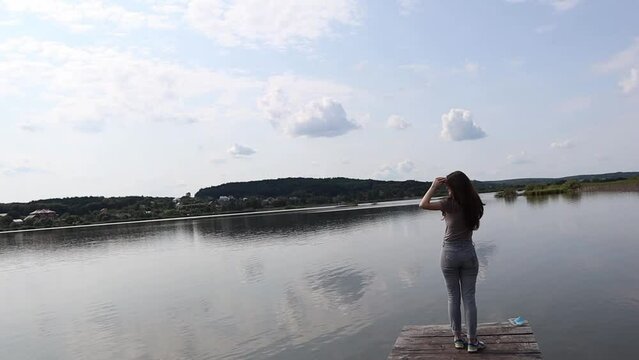 A young woman from behind is standing on a wooden bridge and looking at the lake on a sunny day