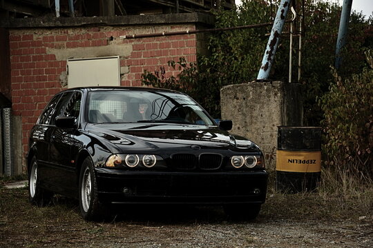 Front eines schwarzen BMW e39 Touring Kombi mit Angel-Eyes steht neben einem Metallfass