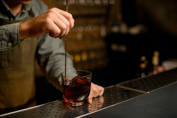 hand of bartender hold long bar spoon and stirring cold drink in glass on bar.