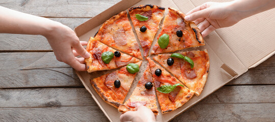 Friends taking pieces of tasty pizza from cardboard box on wooden background, top view