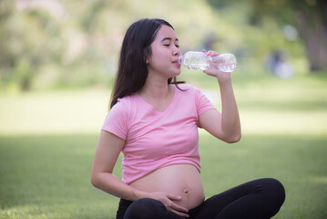 Happy pregnant Asian woman sitting on lawn and drinking water. Pregnancy, motherhood, people and expectations.