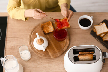 Mature woman making tasty toasts with jam and drinking coffee in kitchen
