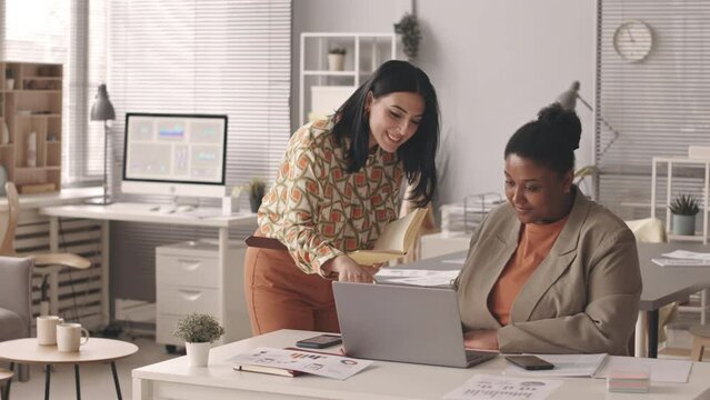 Medium slowmo of two young multiethnic female colleagues looking at laptop screen and having conversation, working together in modern office