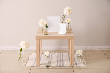 Vases with beautiful peony flowers and blank photo frames on wooden table near light wall in room