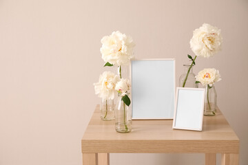 Vases with beautiful peony flowers and blank photo frames on wooden table near light wall