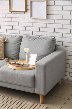 Tray with peony flower, blank photo frame and magazine on sofa near white brick wall