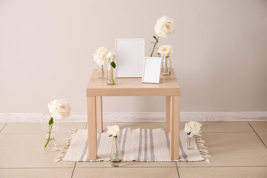 Vases with beautiful peony flowers and blank photo frames on wooden table near light wall in room