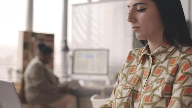 Tilt down medium slowmo of young Middle Eastern woman in patterned shirt typing on laptop at office desk