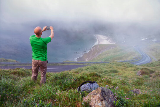 Hiker with backpack taking picture on his smart phone of stunning nature scenery. Keem bay and beach in fog. Travel and holiday concept. Model bald in green shirt. Achill island, Ireland.