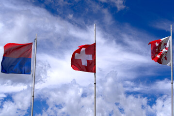 Swiss flag and flag of Canton Ticino and Canton Valais blowing in the wind at summit of Swiss mountain pass Nufenen on a sunny summer day. Photo taken July 3rd, 2022, Nufenen Pass, Switzerland.