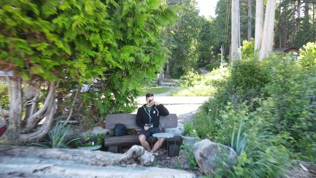 Young man sitting on a bench drinking coffee right at a pebble beach on Canada's sunshine coast. Aerial pull away shot
