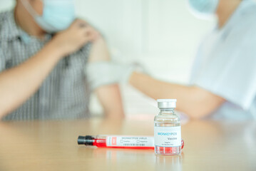 A vaccine vial for monkeypox with the blurry background of a doctor injecting a vaccine.