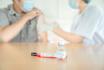 A vaccine vial for monkeypox with the blurry background of a doctor injecting a vaccine.
