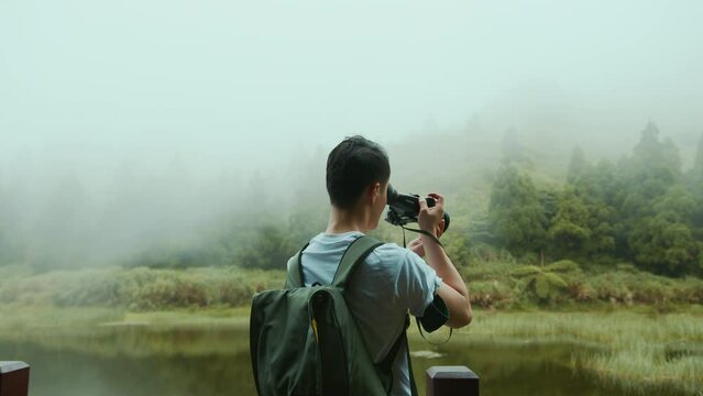 A young Asian male photographer in a white T-shirt and a backpack is taking photographs of nature by a lake in the mountains, in Taiwan. Shot with URSA Mini Pro 4.6K G2