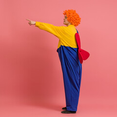 funny clown in a wig and a yellow-blue suit with a propeller on his back stands sideways and points his finger to the side on a colored background