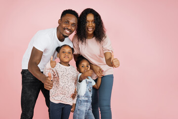 Happy family concept. Young parents and their daughters hugging sitting down on the floor showing thumbs up over pink studio background, free space.