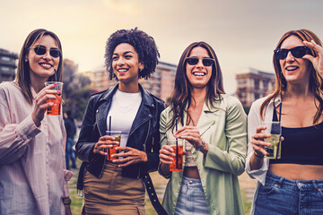 Group of four girlfriends standing outdoors carefree and drinking cocktails - happy young women...