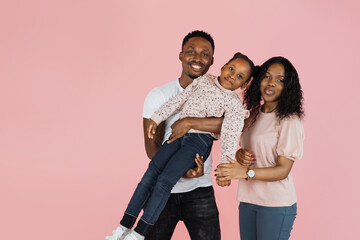 Fototapeta premium Cheerful african man and his wife holding excited daughter on hands. Woman and her husband looking and posing at camera, standing isolated over pink studio background, panorama, banner free copy space