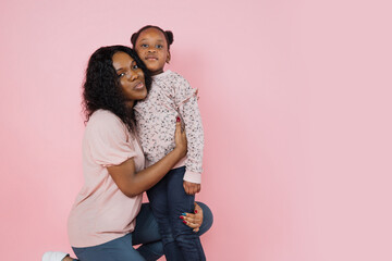 Indoor studio shot of pleasant young African woman mother and her cute adorable little daughter, wearing bright trendy casual clothes, posing together on pink background.