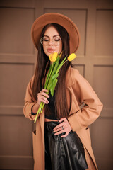 A beautiful stylish brunette in round glasses with makeup in a brown hat and raincoat stands indoors and holds yellow flowers tulips in her arms