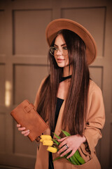 A beautiful stylish brunette in round glasses with makeup in a brown hat and a raincoat stands indoors and holds a brown notebook in her hands and yellow tulip flowers stick out in the pocket of the r