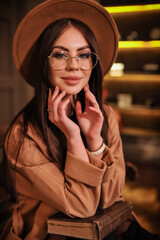 Portrait of a beautiful stylish brunette woman in round glasses with makeup in a brown hat and raincoat sits indoors and touches her face