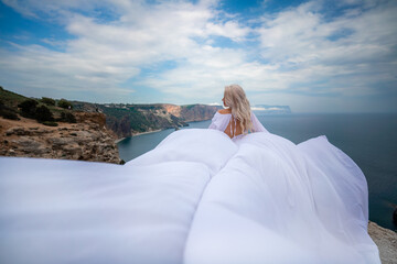 Blonde with long hair on a sunny seashore in a white flowing dress, rear view, silk fabric waving in the wind. Against the backdrop of the blue sky and mountains on the seashore.