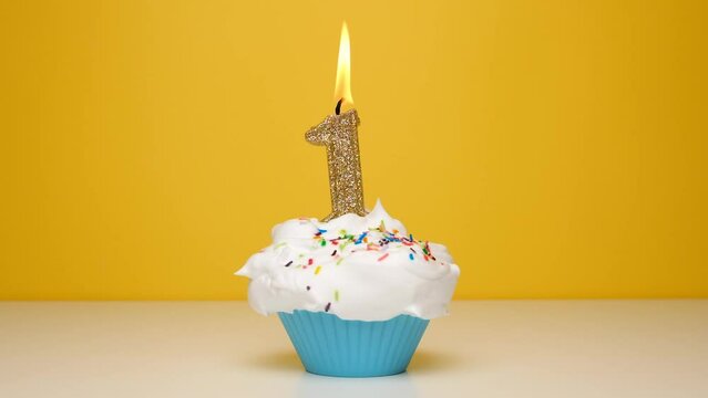 Birthday cupcake With Burning golden Candle number one on bright yellow Background. Slow motion first anniversary video banner.