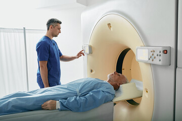 Radiographer operating computed tomography scanner being used to scan lying patient in radiology department of hospital. CT scanner - obrazy, fototapety, plakaty