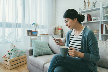 beautiful Taiwanese lady sitting with folded legs on sofa is reading online news on mobile phone...