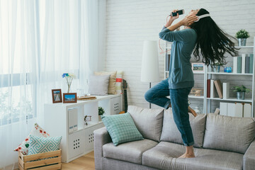 full length asian woman wearing earphones and singing into her phone as a microphone is dancing jumping wildly on the sofa while partying alone at home