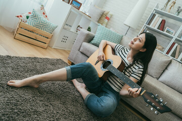 dutch angle shot of pretty asian woman is sitting on floor by sofa and playing an acoustic guitar...