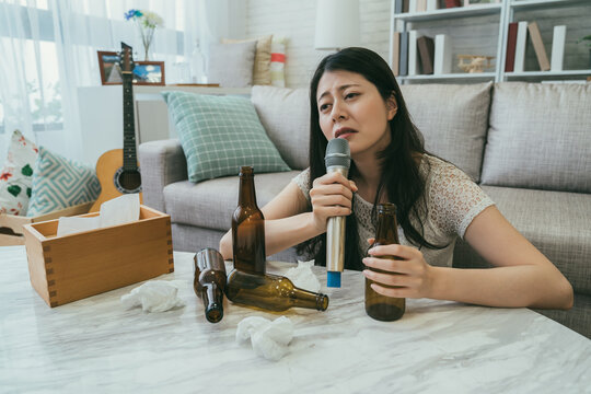 heartbroken asian Japanese woman sitting by a table with beer bottles and snot wads is abusing alcohol while crying singing karaoke alone at home after breakup.