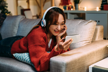 carefree asian girl lying prone on sofa and propping her face is enjoying watching shows through streaming service on her phone in a comfortable home interior.