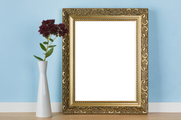 Vertical golden picture frame leaning on blue wall with stonecrop flowers in vase transparent...