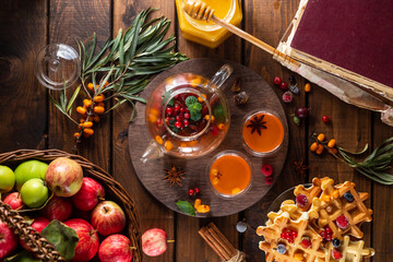 Obraz na płótnie Canvas Autumn tea with berries and honey. Sea buckthorn and raspberries in a glass teapot, cups with poured tea. Apples and waffles with honey.