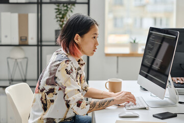 Asian young girl sitting at her workplace in front of computer monitor and typing codes working at...