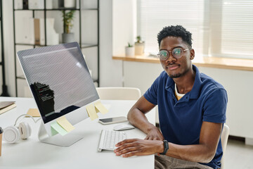 Portrait of African young developer in eyeglasses looking at camera while sitting at his workplace with computer monitor with codes on screen