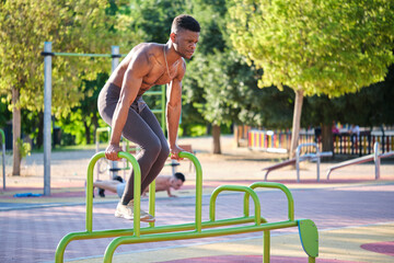 Young fit shirtless black man doing calisthenics workout on parallel bars outdoors on sunny day....