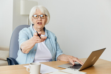 a surprised elderly woman is sitting at her desk at home with a laptop and with her mouth wide open stretches out her hand to the camera
